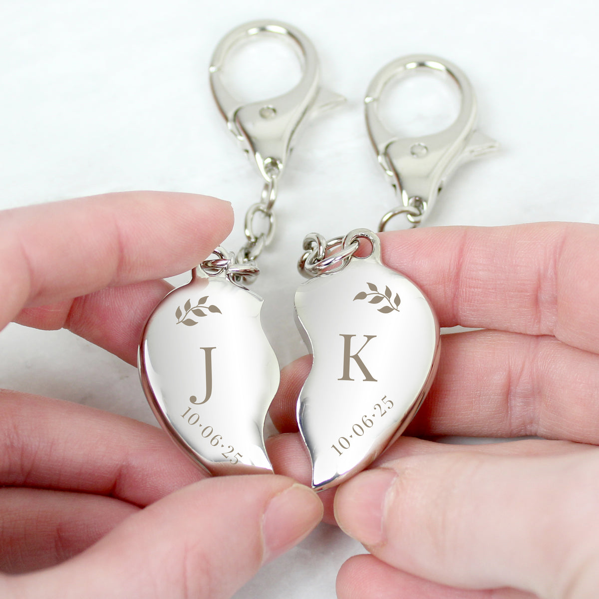 Two silver heart-shaped keychains with initials and date held by hands on a white background