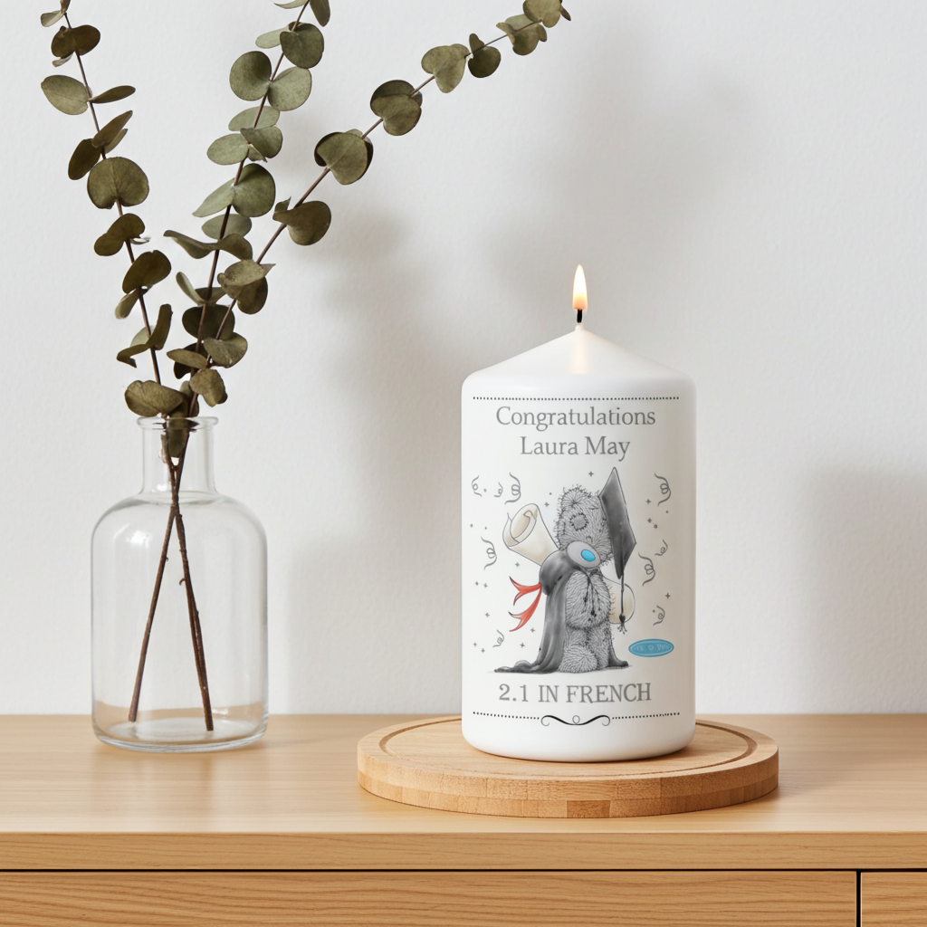 A lit white pillar candle sits on a round wooden coaster on a light wood table, next to a glass vase with eucalyptus branches. The candle is personalized with the text 'Congratulations Laura May' and '2.