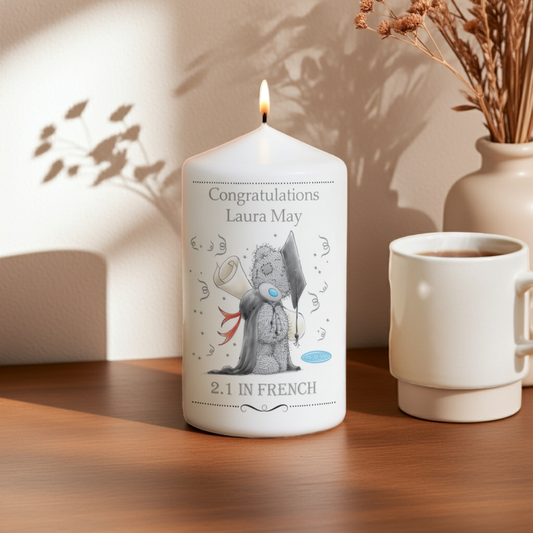 A white taper candle with a lit flame sits on a wooden table, printed with a cute teddy bear illustration and the text 'Congratulations Laura May' and '2.1 IN FRENCH'.