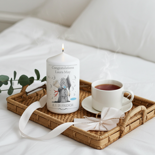A personalized white candle with a lit wick sits on a woven tray beside a cup of tea and a white ribbon, set on a bed with soft white bedding in the background.