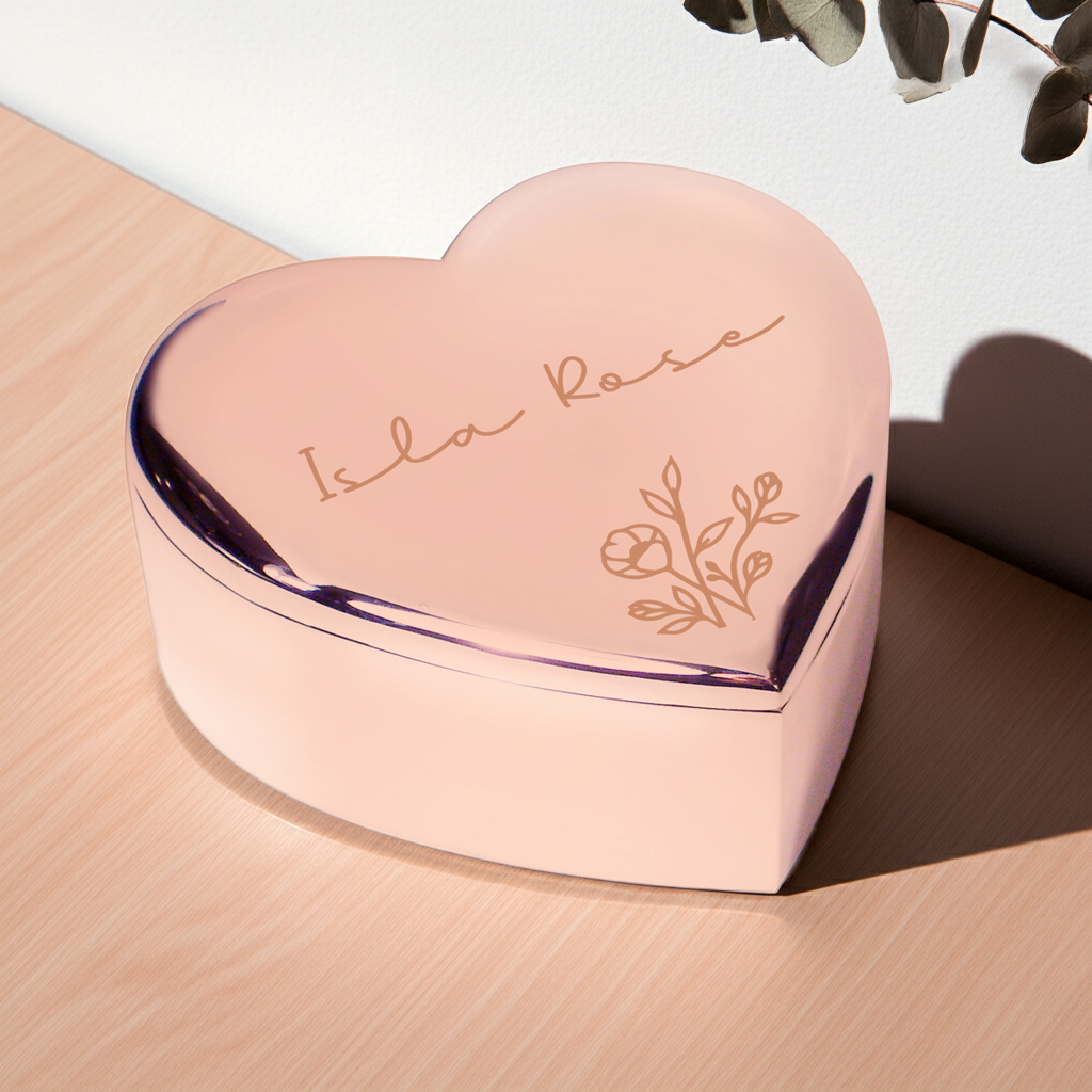 A heart-shaped pink keepsake box with a glossy finish, featuring the engraved text 'Isla Rose' and a floral design on the lid, resting on a light wood surface.