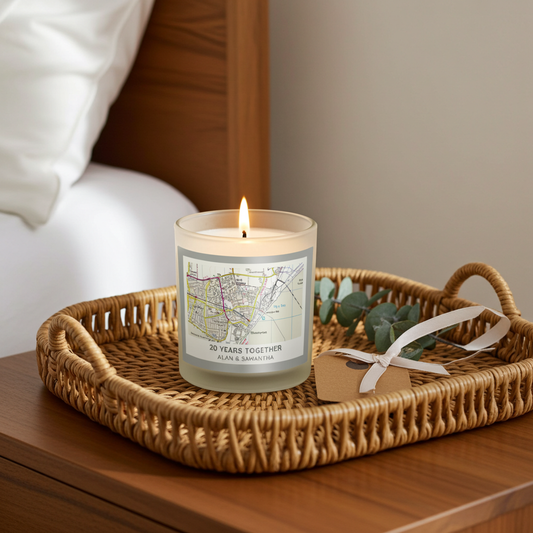 A lit glass candle sits on a woven rattan tray on a wooden bedside table, with a bed and neutral wall in the background. The candle label reads '30 YEARS TOGETHER' and includes a map-style graphic.