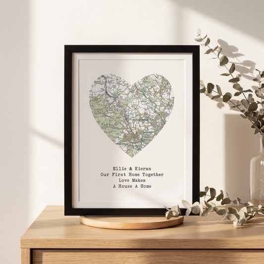 A framed personalized map print featuring a heart-shaped map with the text 'Ellie & Kieran Our First Home Together Love Makes A House A Home' displayed on a wooden stand in a styled interior setting.