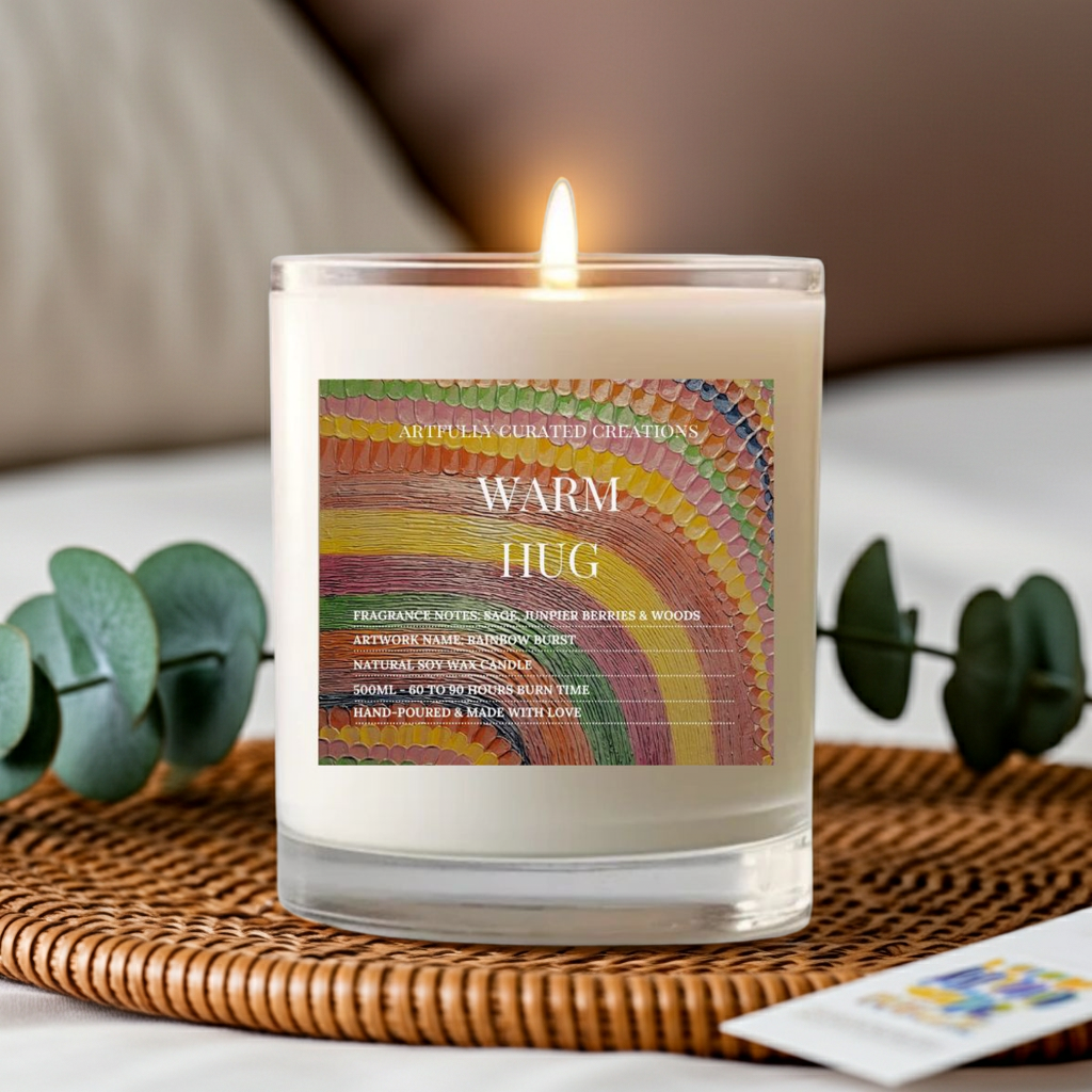 A lit white scented candle in a clear glass jar sits on a woven tray, with a colorful rainbow-style label reading 'WARM HUG' and 'Artful Curated Creations.' The scene is styled with eucalyptus leaves and a soft indoor background.