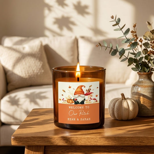 A lit amber glass candle in a round wooden holder sits on a stone surface with eucalyptus branches in the background. The candle label features a fall gnome illustration and the text 'WELCOME TO Our Patch RYAN & SARAH'.