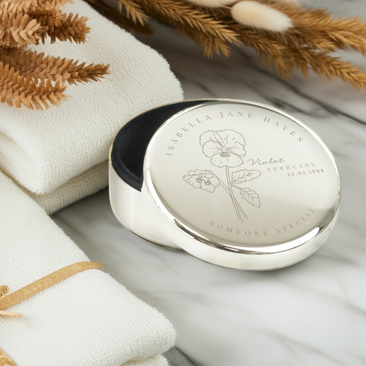 A round silver keepsake box with a polished lid engraved with a flower illustration and the text 'ISABELLA JANE HAYES Violet FEBRUARY 23-02-1998' and 'SOMEONE SPECIAL', styled on a white marble surface with folded white towels and dried botanicals.