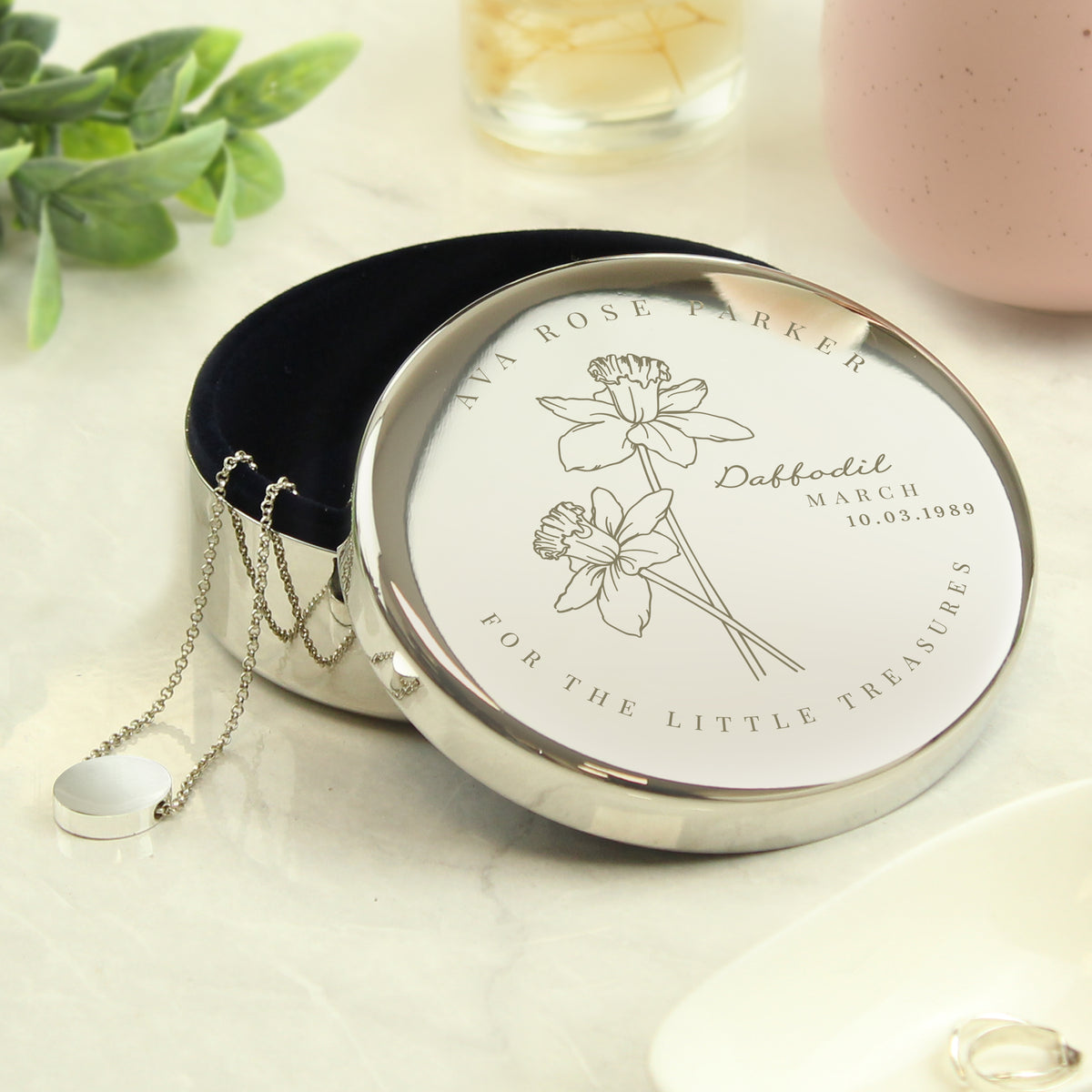 Silver jewelry box with floral design and text on a light surface with greenery.