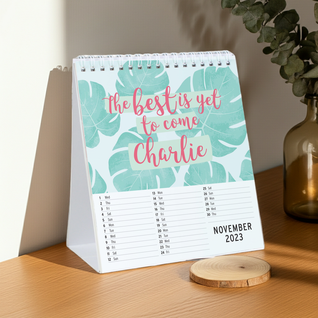 A spiral-bound desk calendar standing on a wooden table, printed with large mint green tropical leaves and pink script text reading 'the best is yet to come Charlie.' The lower section shows the November 2023 calendar dates.