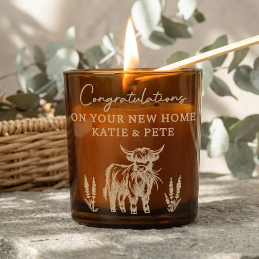 A lit amber glass candle with a wooden match resting in it, printed with the message 'Congratulations ON YOUR NEW HOME KATIE & PETE' and a cow illustration, set on a stone surface with blurred greenery and a wicker basket in the background.