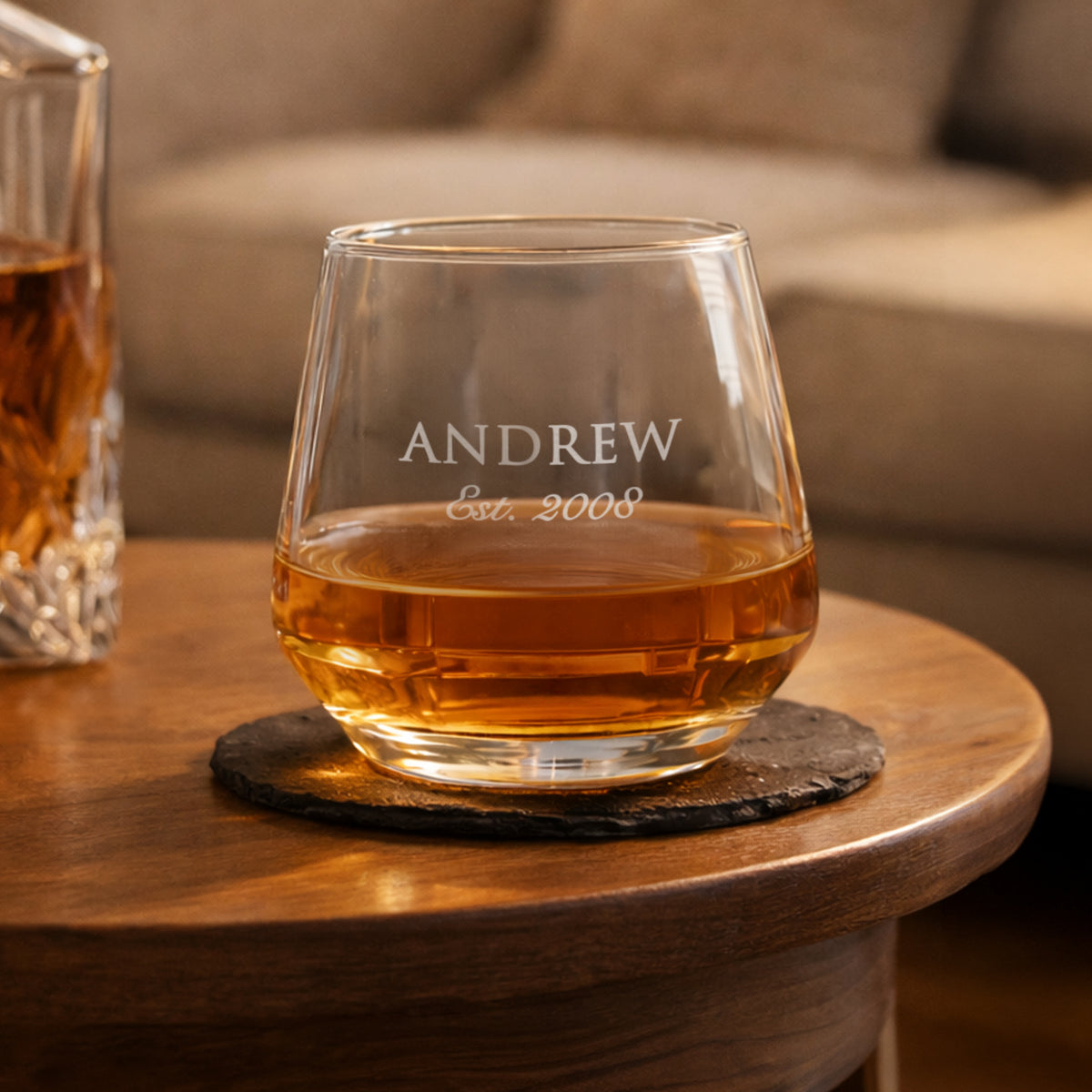 Personalized whiskey glass with 'ANDREW Est. 2008' on a wooden coaster with a blurred background.