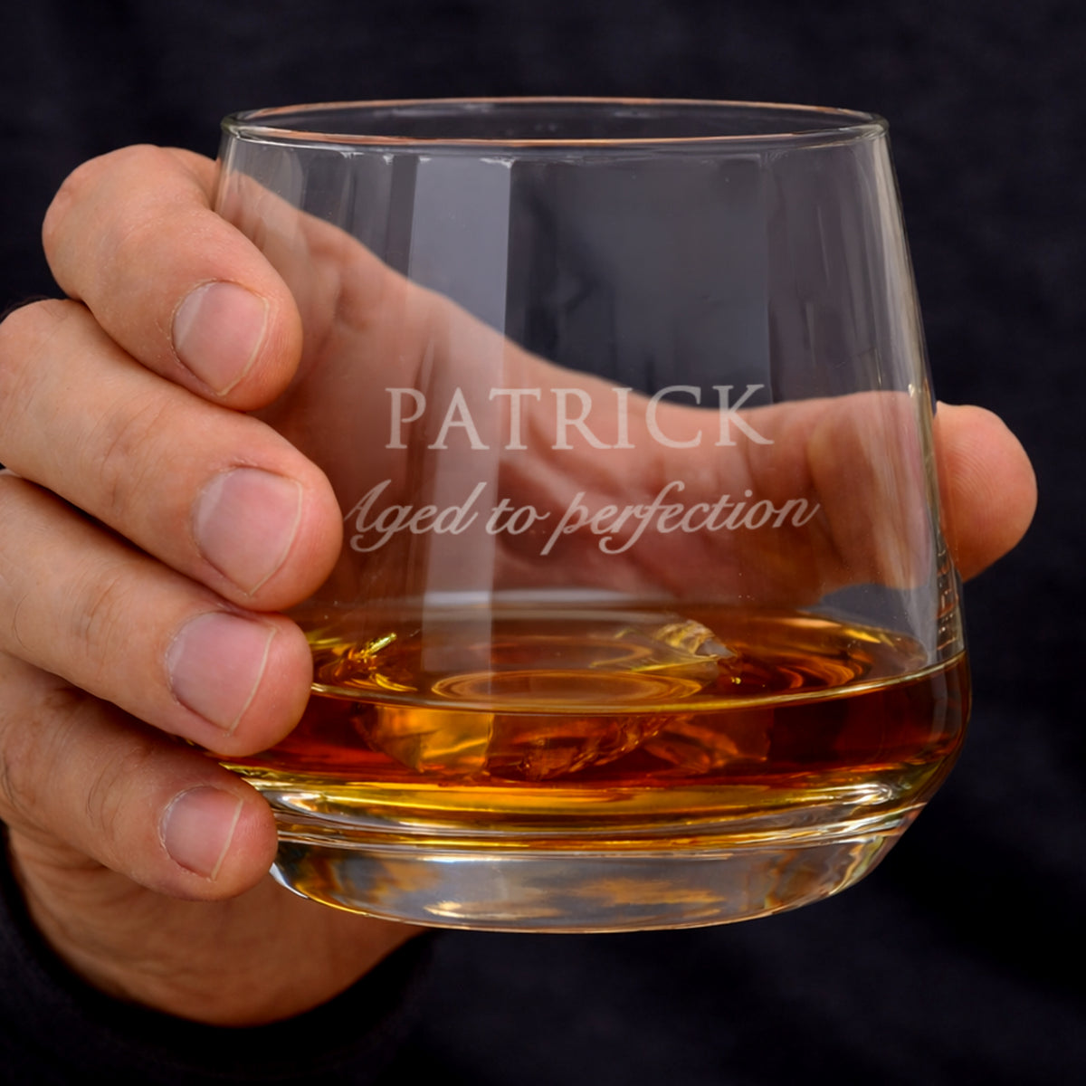 Hand holding a glass of whiskey with 'Patrick aged to perfection' engraving.